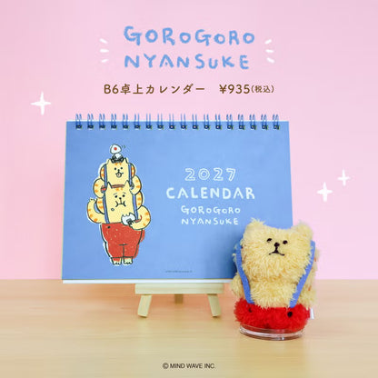 A 2027 desk calendar titled 'GOROGORO NYANSUKE' displayed on a small easel stand, with a plush teddy bear accessory in front. The scene is set against a pink background with Japanese text and a light wood tabletop.