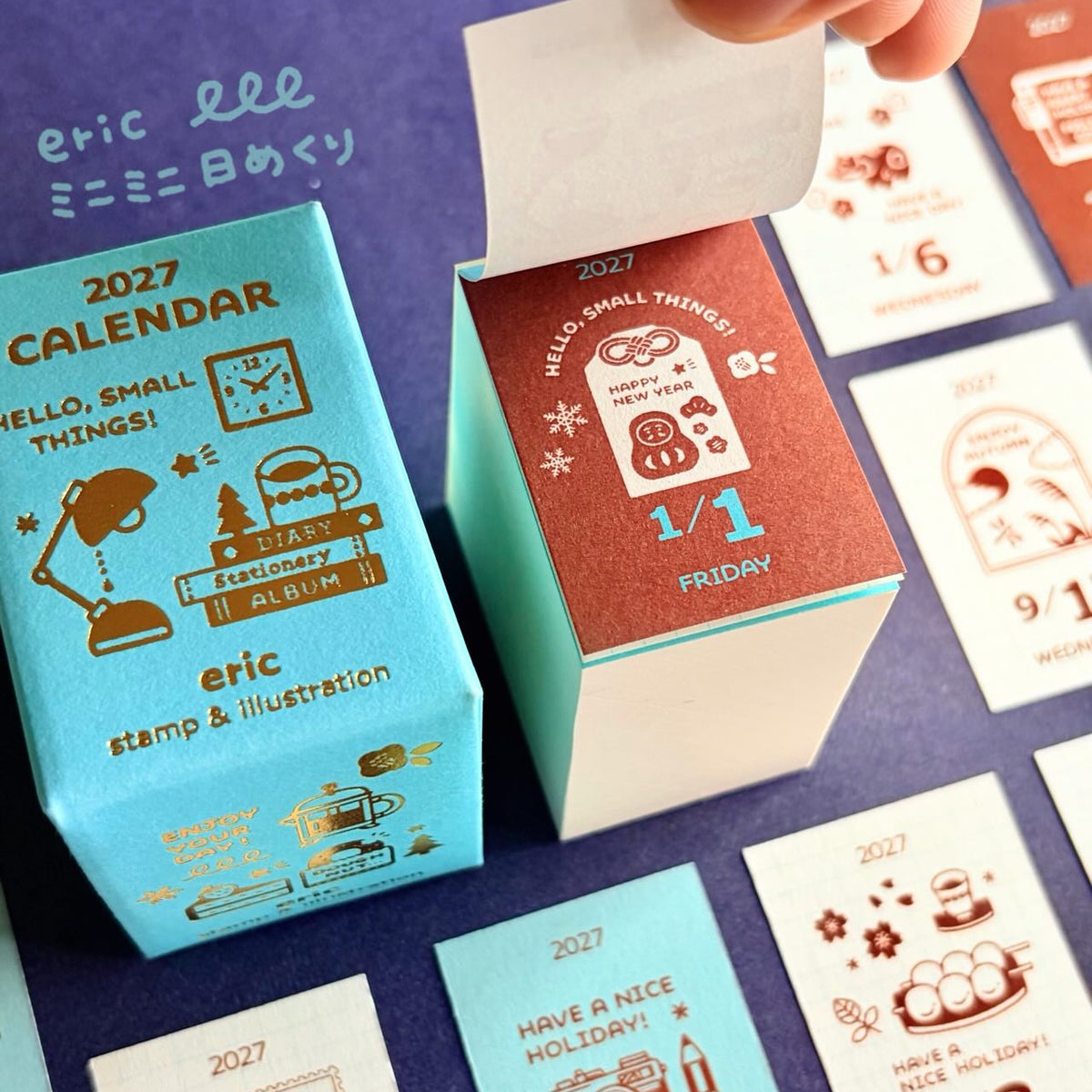 A close-up of a 2027 daily calendar and its decorative boxes on a dark purple surface. A hand is lifting the lid of one coral-red box, while other cards and boxes in light blue and white are arranged around it.