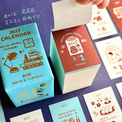 A close-up of a 2027 daily calendar and its decorative boxes on a dark purple surface. A hand is lifting the lid of one coral-red box, while other cards and boxes in light blue and white are arranged around it.