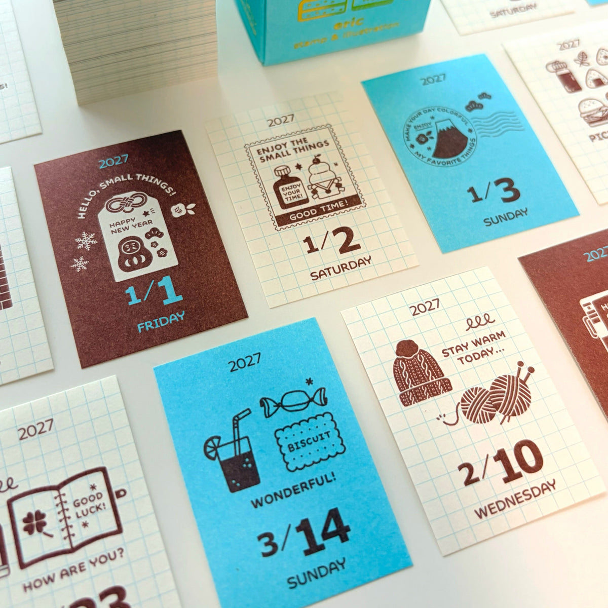 A close-up of a printed daily planning or card deck set with multiple square cards laid out on a white surface and a turquoise box standing behind them. The cards feature different colors including turquoise, dark brown, cream, and light blue, with illustrated icons and text such as dates, days of the week, and short phrases.