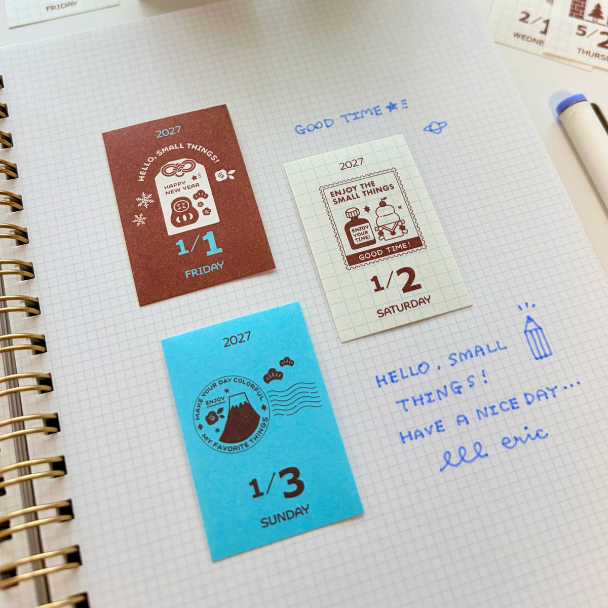 A close-up of a spiral notebook page with several small printed sticker or label cards arranged on the paper, along with handwritten notes and a pen. The cards show dates and illustrations in brown, cream, and bright blue, and one card is being held by a hand at the top edge.